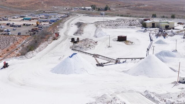 Salt mining site in Nevada showing white salt mounds, conveyor belts, storage buildings, and vehicles moving across an arid reddish brown landscape.