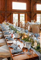 Rustic wedding reception table set with elegant blue glassware, candles, greenery, and floral arrangements, creating a warm and inviting atmosphere in a wooden barn venue