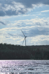 Wind turbine on a lake with forest and reflection under a partly cloudy sky.