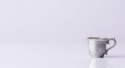 Ornate silver cup with intricate patterns on a reflective white surface, showcasing detailed craftsmanship and elegant design.