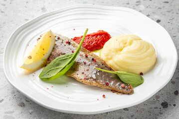Cooked fish fillet with lemon and spinach, creamy mashed potatoes, and red sauce on a white plate. Gourmet meal on gray countertop, ideal for food photography and culinary presentation