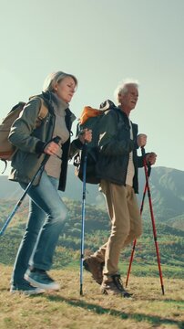 Vertical slow motion video senior couple trekking with backpacks and poles in mountains summer travel adventure lifestyle active holiday freedom healthy aging nature scenic landscape outdoors