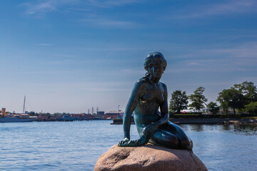 Naklejka premium Copenhagen, Denmark 07-20-2025 The bronze statue of the Little Mermaid sitting on a rock by the waterside at the Langelinie promenade in Copenhagen. The small and unimposing statue is an icon in town.