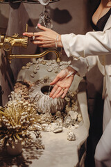 Young woman with stylish pearl beads washes hands at seashell-decorated washbasin indoors