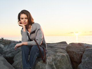 A thoughtful woman sits on rocky shore at sunset, resting her chin on her hand. She wears a gray...
