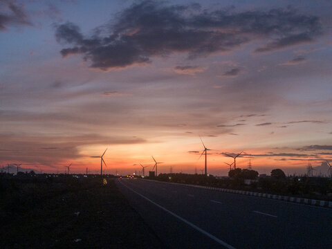 A scenic evening highway view with silhouettes of wind turbines against a dramatic sunset sky, blending warm orange and purple hues, symbolizing renewable energy and peaceful travel moments