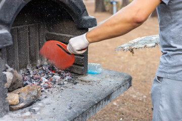 A young Caucasian man with short hair uses a red shovel to remove ashes from a stone barbecue grill...