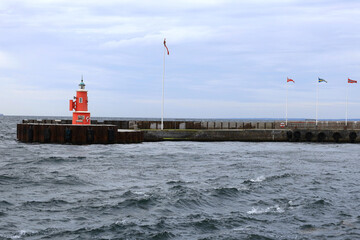 Landscape photo with a view of a red lighthouse, flags of Denmark, Sweden and Norway on flagpoles and the sea with waves during a light storm early in the morning in the city of Helsingor, Denmark