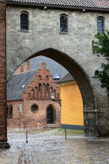 Photo with a view of a stone arch and a red brick building on the grounds of Roskilde Domkirke Cathedral during the rain in Roskilde, Denmark