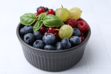Different ripe juicy berries and basil in bowl on white tiled table, closeup