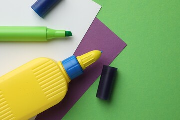 Glue in bottle, felt pen and color paper on green background, flat lay. Space for text