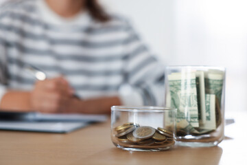 Glass jars with money and woman budgeting at wooden desk, selective focus