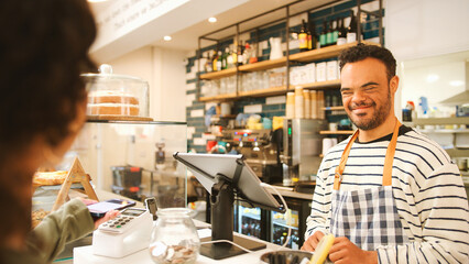 Fototapeta premium Man With Down Syndrome Serving Female Customer At Coffee Shop Checkout Using Contactless Payment