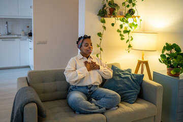 Calm black woman sitting on sofa in living room, practicing deep breathing exercise with hands on chest, promoting relaxation, mindfulness. Serene focused african american female meditating at home. 