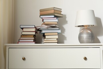 Many stacked books and lamp on white chest of drawers indoors