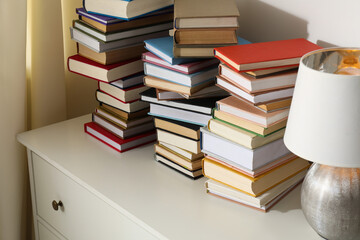 Many stacked books and lamp on white chest of drawers indoors, closeup