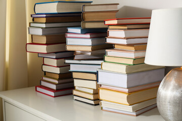 Many stacked books and lamp on white chest of drawers indoors, closeup