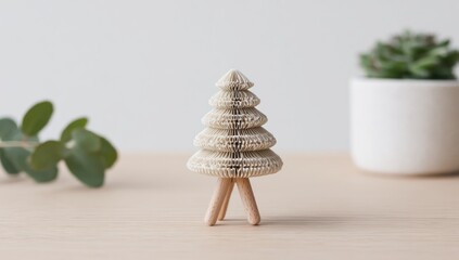 A delicate, paper-crafted Christmas tree ornament sits atop a light beige wooden surface, accented by soft, muted tones and a touch of greenery.