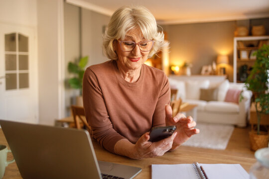 Senior woman with laptop on desk using mobile phone to pay household bills online while sitting at home - Powered by Adobe