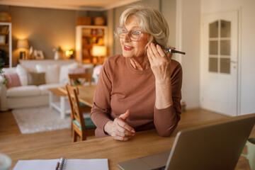 Elderly woman with glasses listening to doctor consultation on smartphone speaker while sitting at desk at home