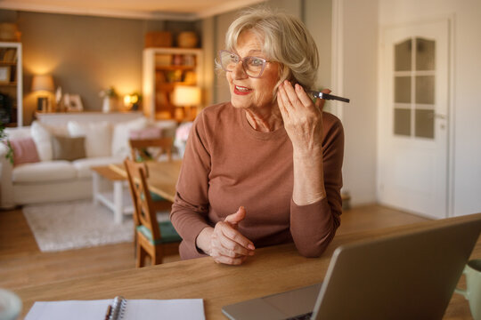 Elderly woman with glasses listening to doctor consultation on smartphone speaker while sitting at desk at home