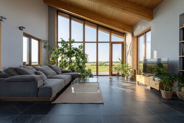 Fototapeta premium A modern living room with large windows showcasing a tranquil countryside view, featuring a gray sectional sofa, a glass coffee table, and natural wood accents.