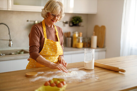 Happy senior woman in eyeglasses kneading dough and preparing homemade sweets on kitchen island at home