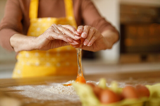 Hands of elderly woman breaking egg into flour on kitchen counter while blogging cookie recipe at her home