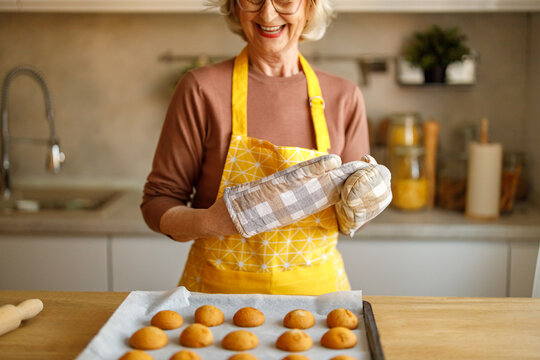 Midsection of happy senior woman wearing oven mitts standing beside freshly baked cookies at kitchen counter