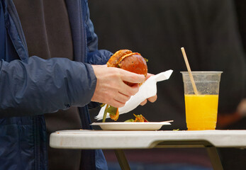 Midsection view of a visitor enjoying a juicy burger with a glass of orange juice at a food festival in Karlin, Prague, authentic street food atmosphere.