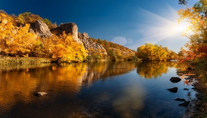 Naklejka premium Golden Autumn Foliage Reflects On Tranquil River Under Bright Sunlight Near Rocky Outcrop