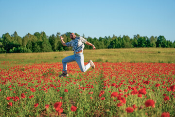 A smiling man jumps with arms outstretched in a poppy field under a bright blue sky. The field is full of red poppies, and trees line the horizon