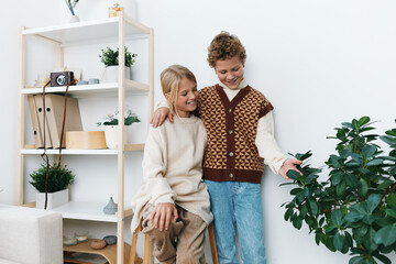 Two joyful children indoors share a warm moment, wearing cozy knit outfits as one points to a lush houseplant in a bright, inviting living space.