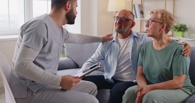 Doctor supports a worried senior couple. A healthcare consultation at home shows empathy, counseling, and support. Nurse or caregiver with a clipboard comforts sick seniors during a sensitive visit.