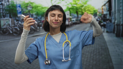 Fototapeta na wymiar Woman nurse wearing blue scrubs and stethoscope raises phone and clenches fist in city street; pride celebration.