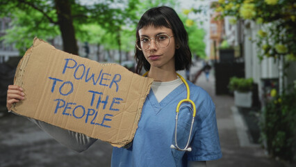 Woman nurse in blue scrubs holds protest cardboard sign reading power to the people on urban street; solidarity.