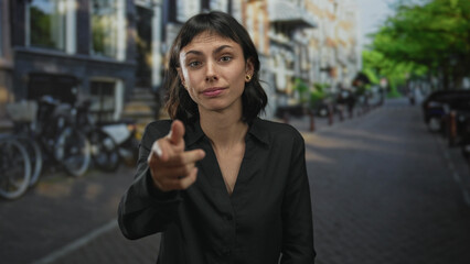 Fototapeta premium Young hispanic woman points finger toward camera with friendly smile on sunlit busy urban street; confidence.