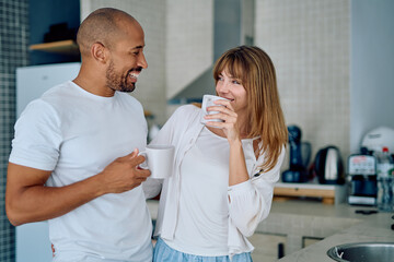 Fototapeta premium Happy interracial heterosexual couple laughing and drinking coffee while standing together in their modern home kitchen, starting the day