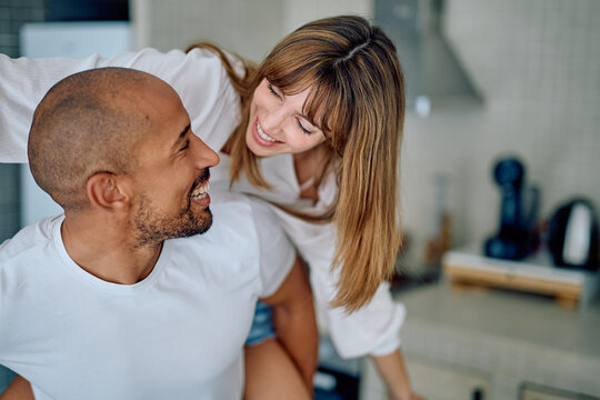 Happy interracial couple piggybacking in home kitchen, smiling and laughing together, showing love and playful relationship