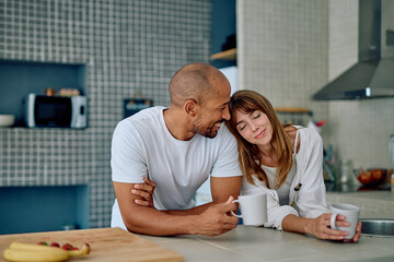 Happy multiracial couple drinking coffee and embracing in a modern kitchen, sharing a loving, peaceful morning moment