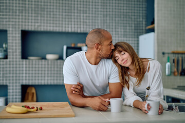Affectionate interracial couple sharing a tender moment, man kissing woman's forehead while holding coffee cups in their kitchen