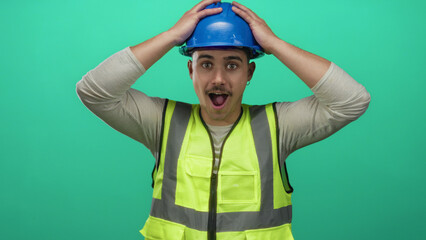 Young hispanic man wearing reflective vest and hardhat stands in front of an isolated green background wall, expressing surprise and holding his head.