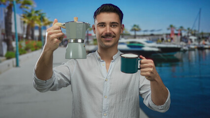 Young hispanic man enjoying coffee while holding a cup and coffee maker by the seaside at a port with boats and palm trees in the background on a sunny day