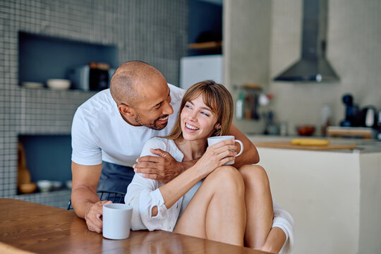 Happy multiracial couple embracing and sharing a laugh while drinking coffee together in their modern kitchen, enjoying a loving morning