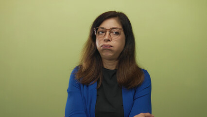 Woman wearing glasses and blue cardigan crosses arms in studio setting against pale green wall;...