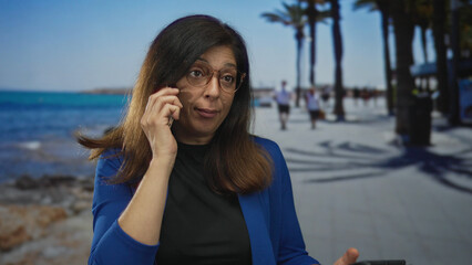 Woman holding phone to ear and tablet while wearing glasses on seaside street; communication focus.