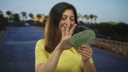 Hispanic middle aged woman holds fan of pesos and pumps fist by green park pathway under sunlit...