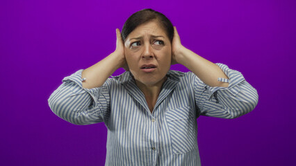 Hispanic woman wearing striped shirt covers ears with both hands in studio under purple lighting;...