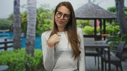 Woman smiling with hand to chest and long hair at hotel building pool deck near palm cabana, casual pose outdoors resort; contentment.