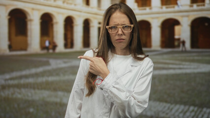 Obraz premium Woman scientist in white lab coat points finger to side at old town building courtyard; confusion concern.